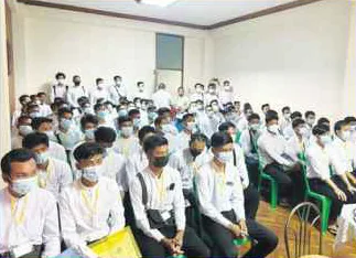A room filled with seated candidates in white shirts, facing the front of the class