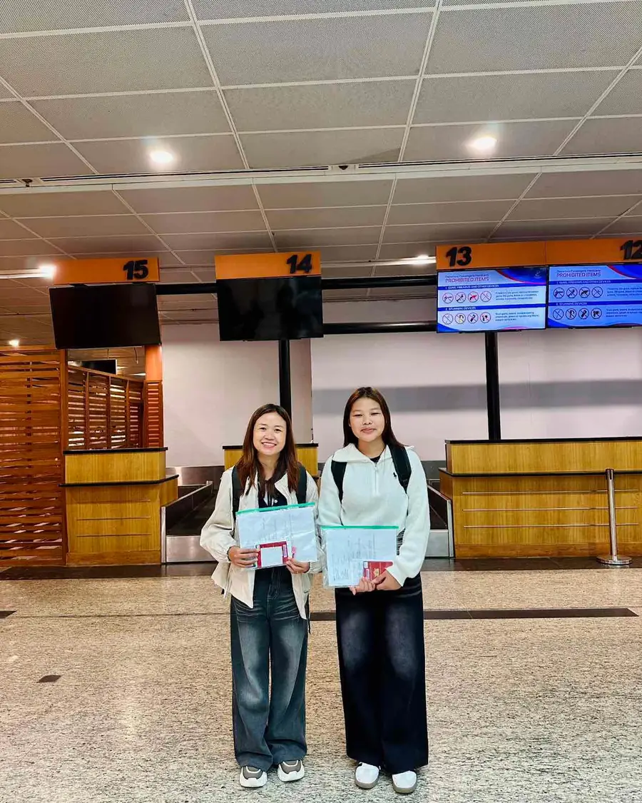Two candidates standing in front of airport departure counters, each holding a document wallet and a passport