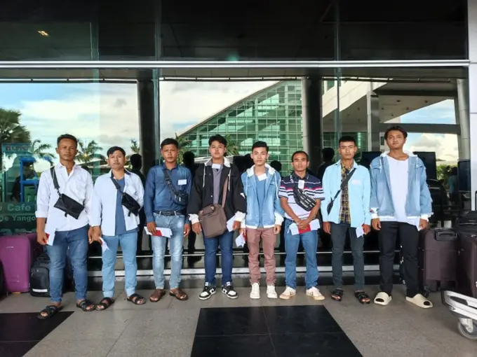 Eight men standing in a line outside a terminal building, several wearing name badges, papers in hand and suitcases beside them