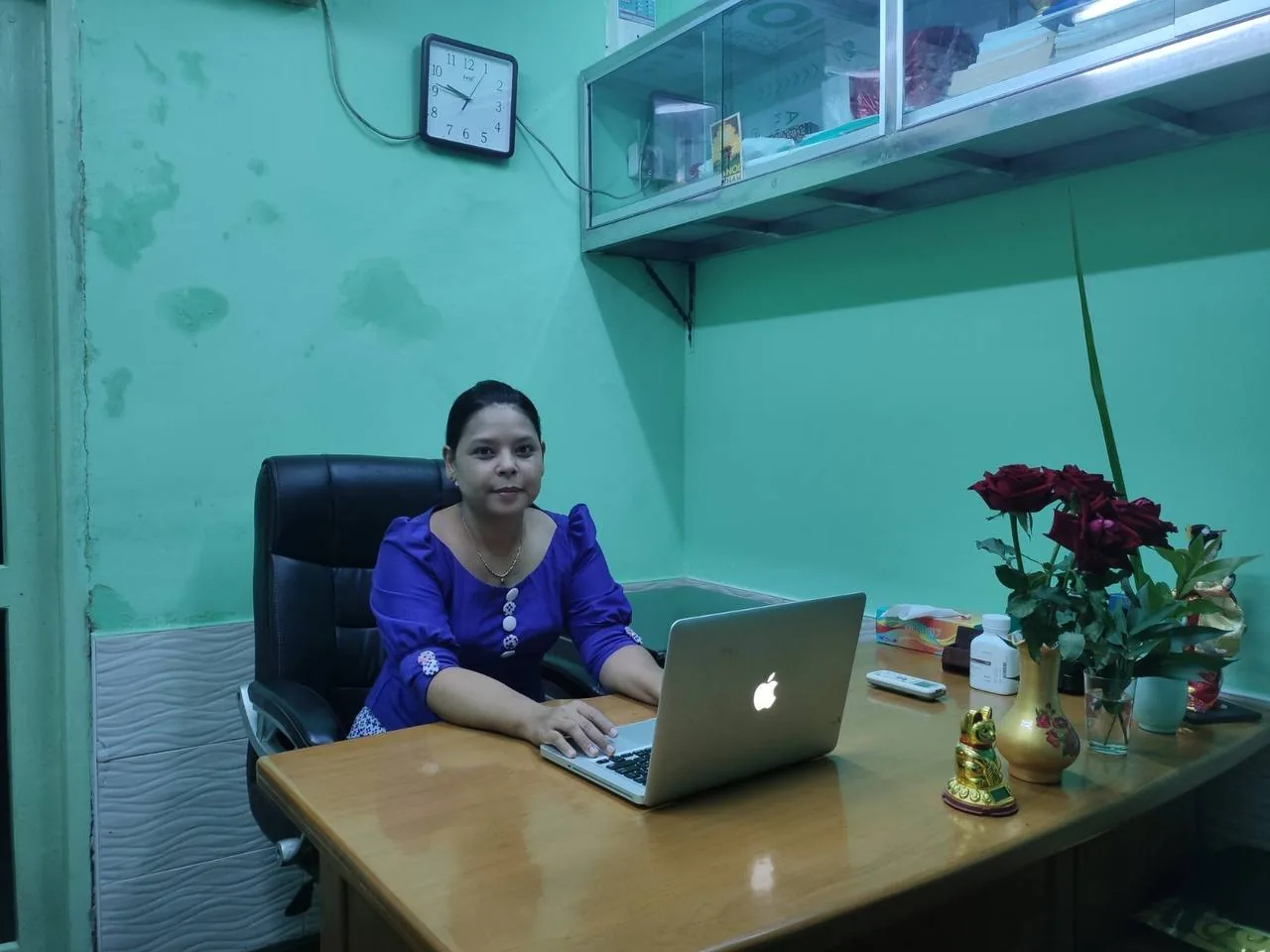 A woman working at a desk with a laptop in the Golden Rain office, a wall clock and a glass-fronted storage shelf behind her