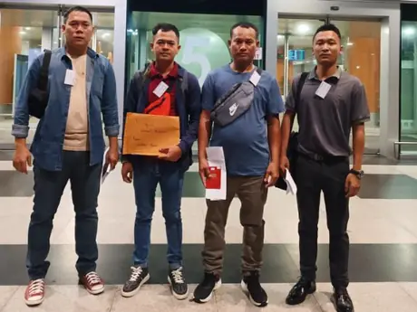 Four men standing outside a terminal door at night, one holding a large envelope, another a passport
