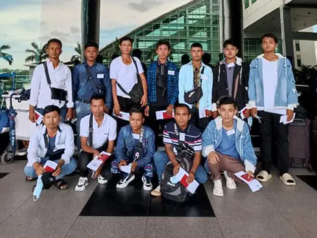 Thirteen men in two rows outside a terminal in daylight, shoulder bags on and papers in hand